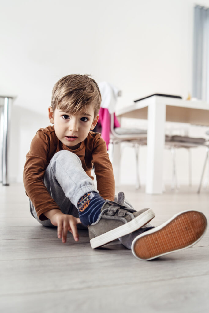 Vedere pe toată lungimea unui băiețel caucazian drăguț, copil mic, care stă pe podeaua de lemn sau de vinil laminat acasă, punându-și sau scoate pantofii în vedere frontală singur
