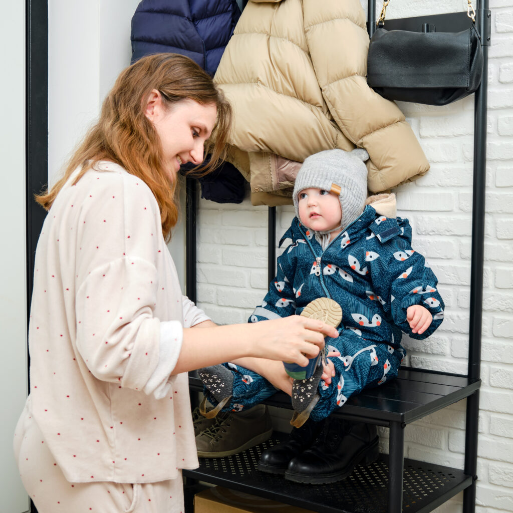 Mama pune un pantofi albaștri pe picior bebeluș care stă pe holul de acasă. Mama femeie îmbrăcă cizme calde haine pe copil pentru plimbare de iarnă pe vreme rece. Copil în vârstă de un an și trei luni
