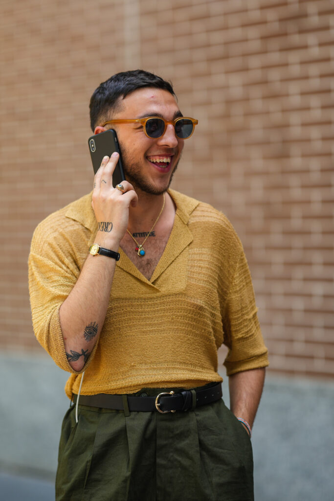 MILAN, ITALY - JUNE 18: A guest wears brown sunglasses, a gold chain pendant necklace, a yellow mustard with embossed striped pattern V-neck polo shirt with long sleeves, a black shiny leather watch, rings, a black matte leather belt, dark green suit pants , outside the Fendi fashion show during Milan Fashion Week Menswear S/S 2023 on June 18, 2022 in Milan, Italy. (Photo by Edward Berthelot/Getty Images)