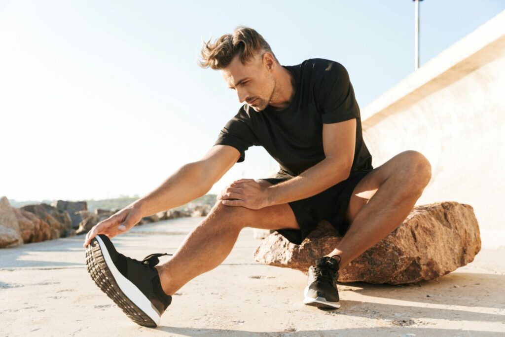 Un bărbat care se întinde înainte de un jogging de dimineață pe plajă într-un tricou negru, pantaloni scurți și pantofi sport pentru bărbați